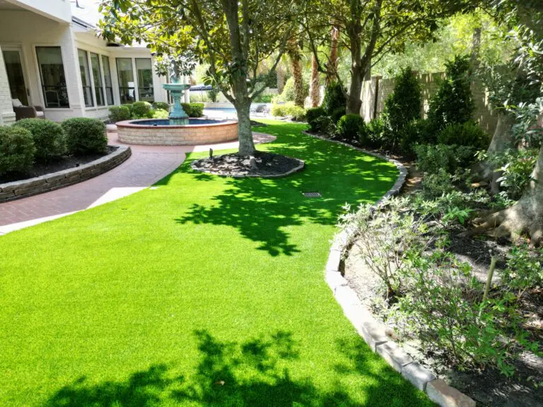 Completed artificial grass lawn around preserved magnolia trees in Friendswood, Texas, with stone-edged garden beds, a fountain and a curved walkway.